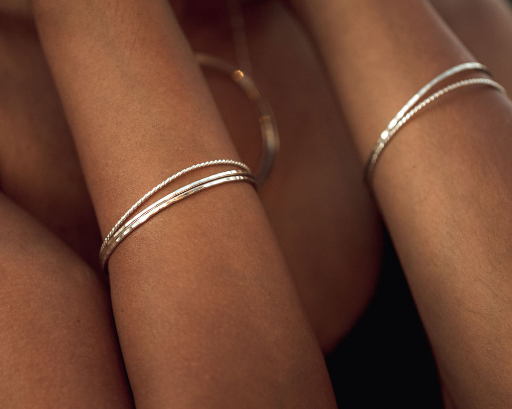 Image shows close up of model wearing several of our handmade sterling silver bangle bracelets. Their smooth, polished texture, delicate chime sound and dynamic dance about your wrists are delightful. A wonderful addition to any ensemble.